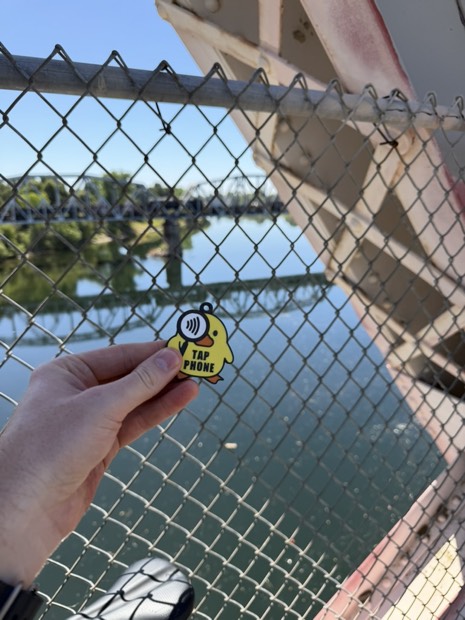 A hand holding a freshly found Where the Duck tag on a bridge over the river